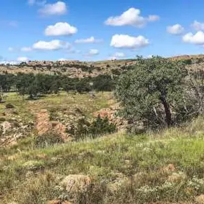 A grassy, rocky Oklahoma landscape with scattered trees and low hills under a blue sky dotted with fluffy white clouds.